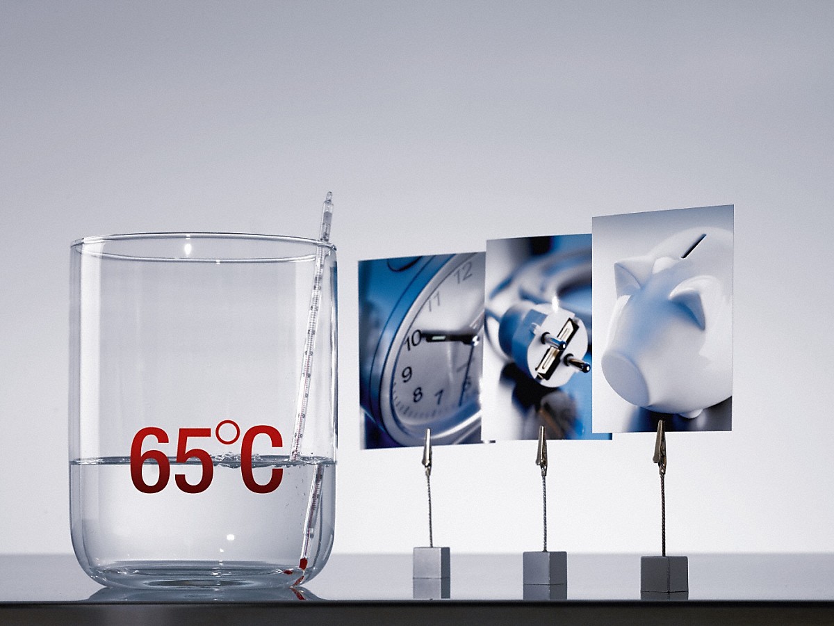 etwas wasser in einem glas mit roter 65°C aufdruck neben drei Fotoklipphaltern mit einer Uhr, einem Steckdosenkabel und einem Sparschwein vor einer hellgrauen Wand