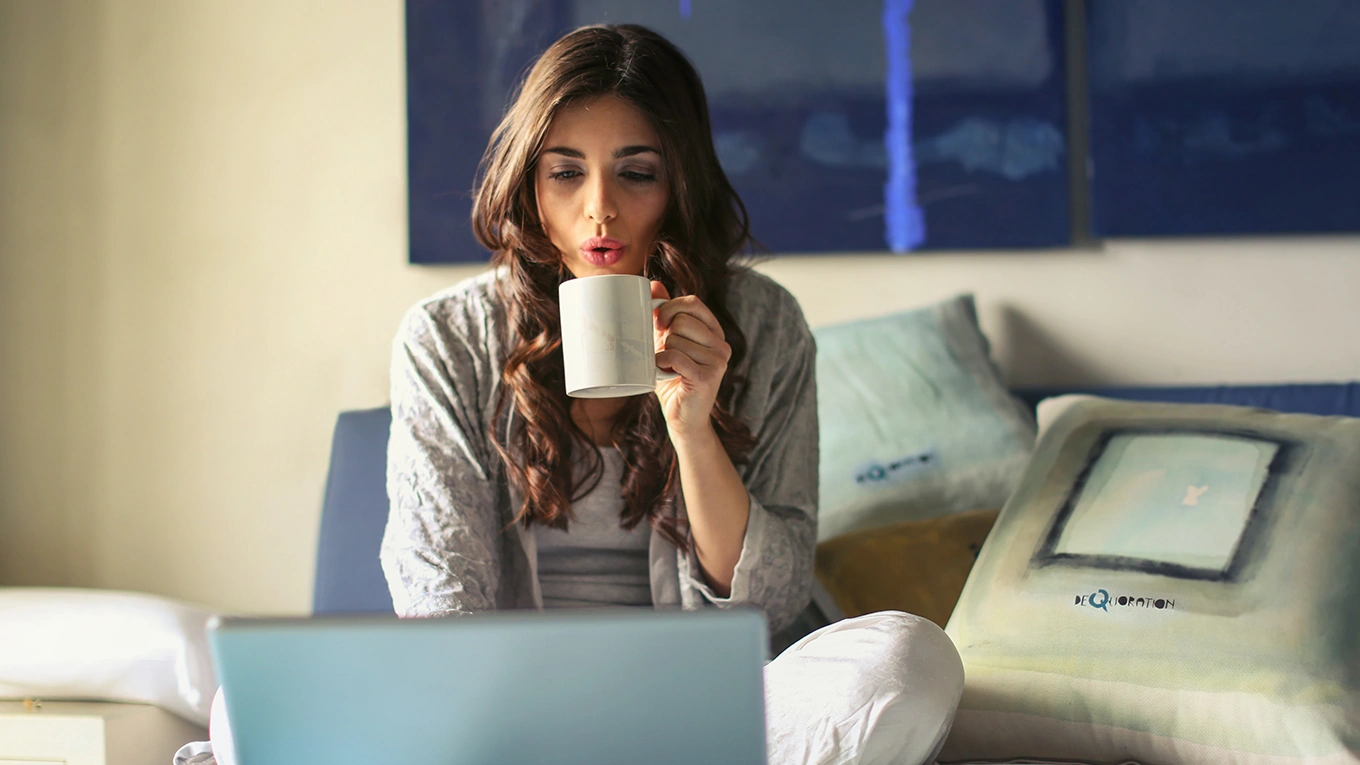 Home Connect auf dem bett sitzende frau die vorm laptop sitzt und in ihre tasse pustet als symbolbild für home connect