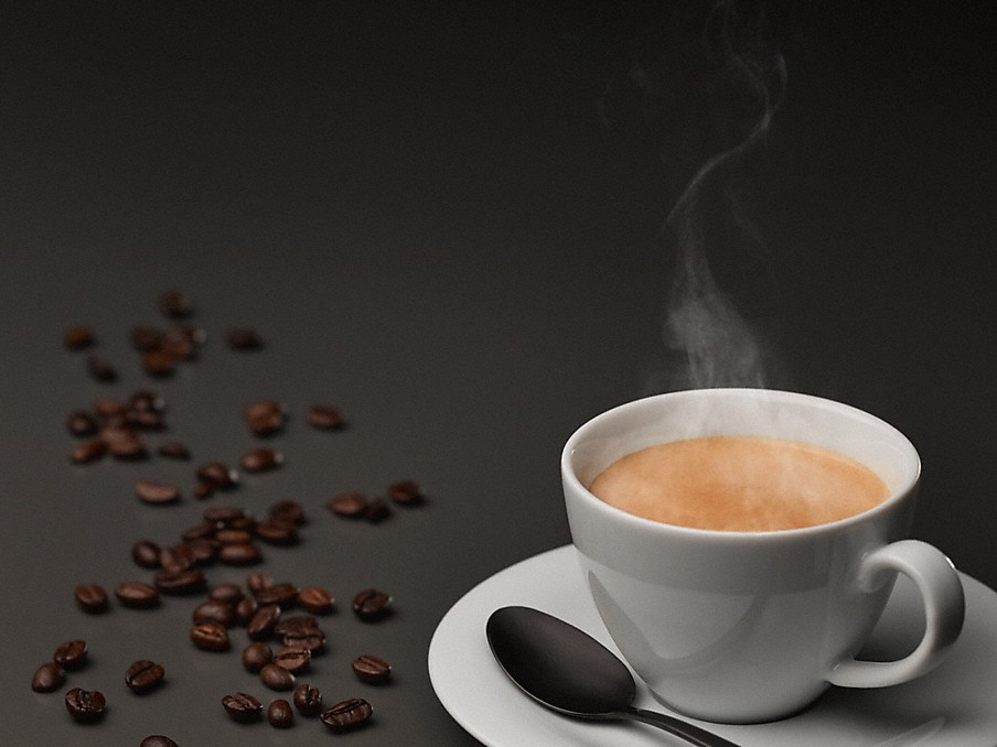 kaffeebohnen auf einer dunklen fläche neben einer weissen Kaffeetasse mit dampfendem Kaffee auf einer untertasse mit loeffel