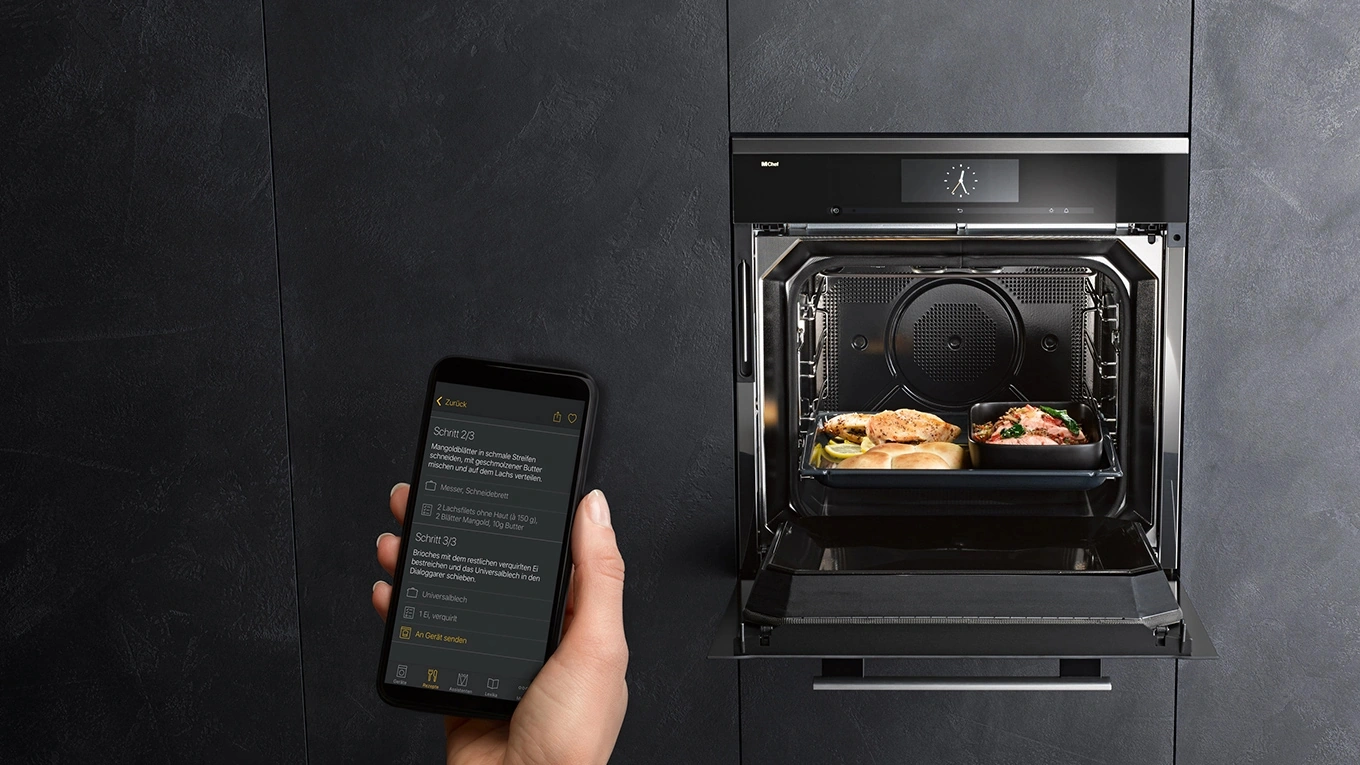miele dialoggarer mit miele@home steuern Miele Home Connect Beispiel mit Smartphone vor einem geöffneten Miele Backofen in einer schwarzen Küchenfront