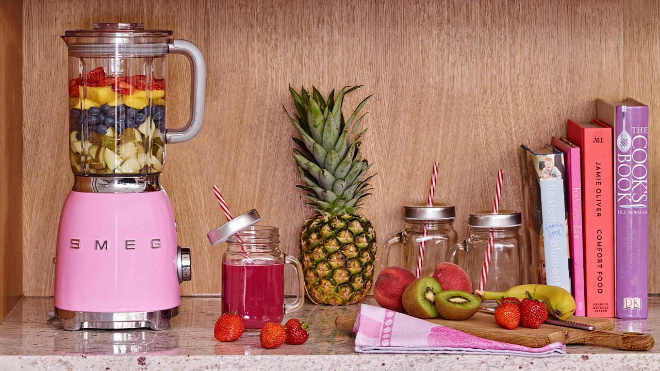 smeg mixer in cadillac pink mixt smoothie und steht im regal neben büchern und früchten